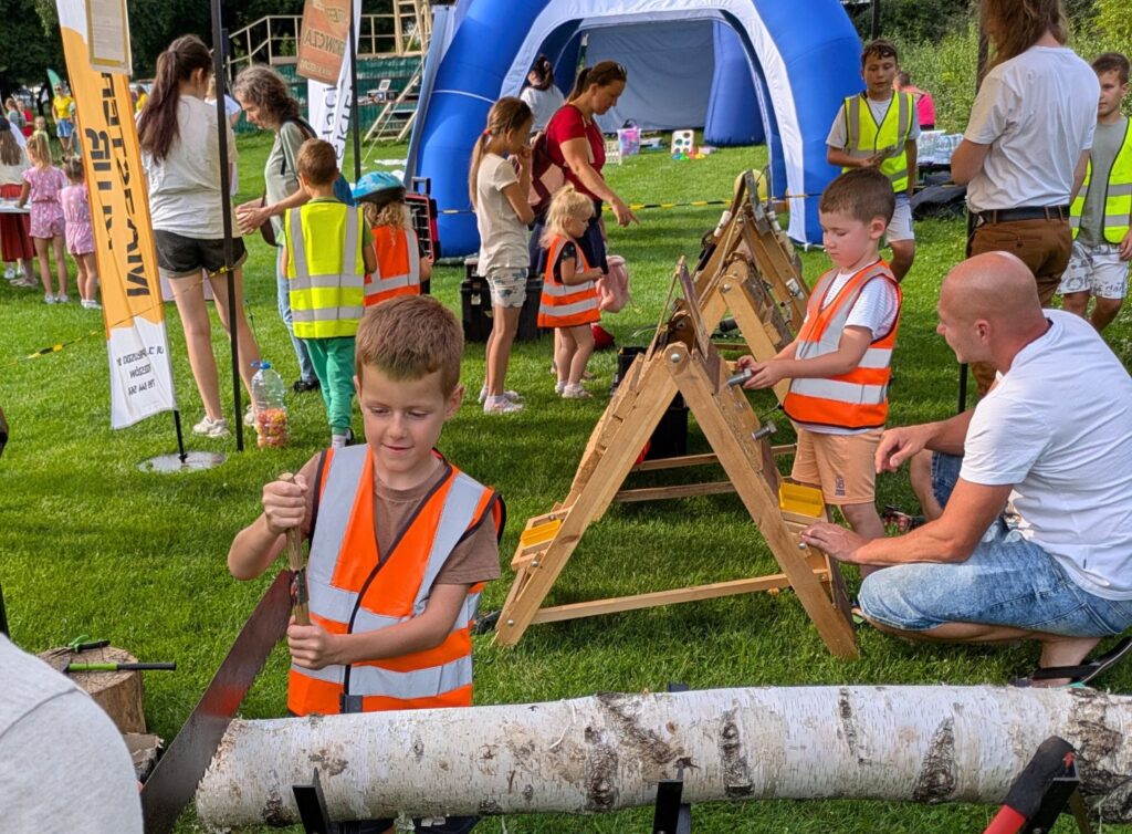Dzieci w kamizelkach odblaskowych uczestniczą w plenerowych warsztatach majsterkowania Fundacji AS KIER, piłując drewno przy pomocy dorosłych.