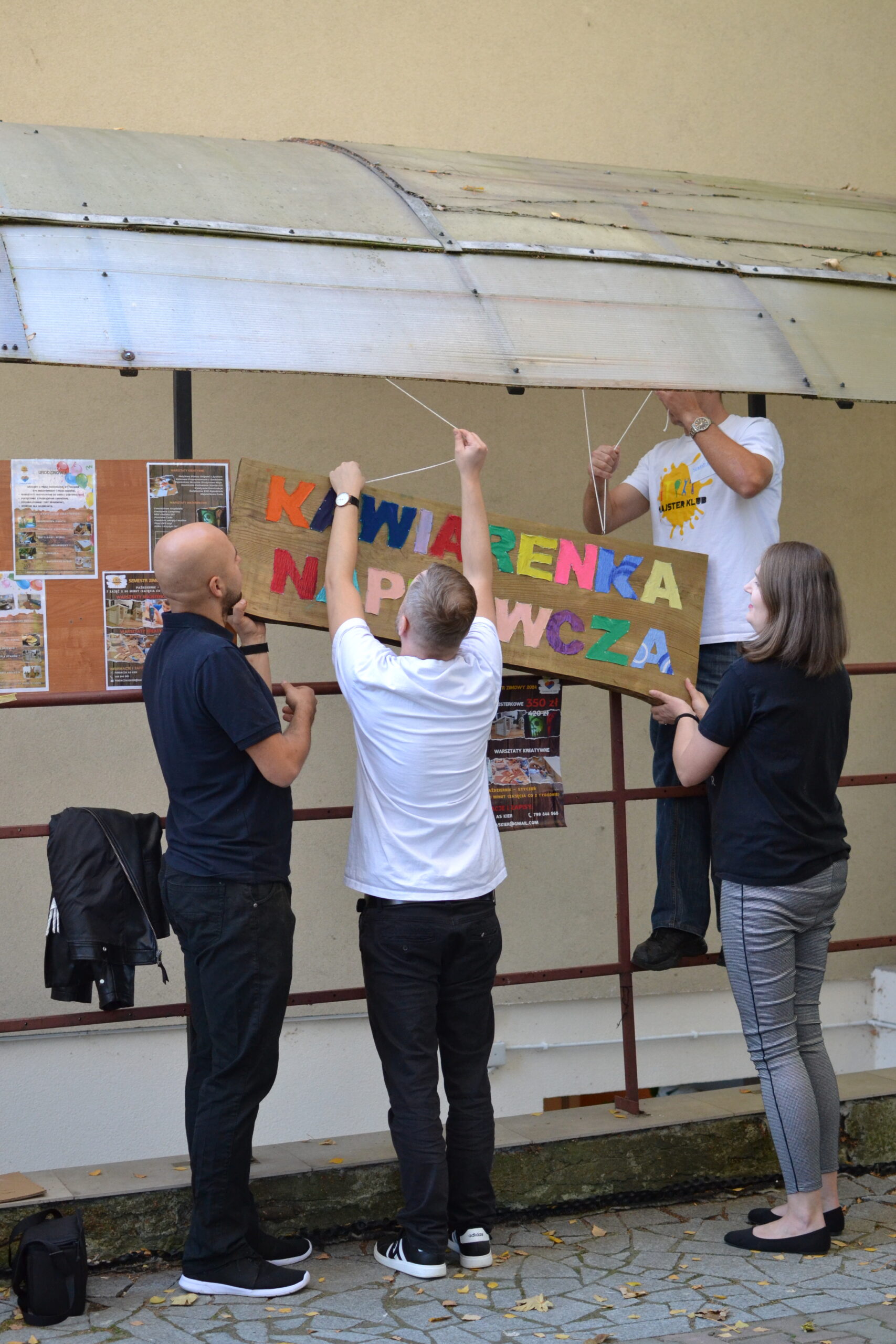 Kawiarenka naprawcza – uczestnicy wspólnie montują ręcznie wykonaną tablicę informacyjną, przygotowując przestrzeń do sąsiedzkich spotkań i działań społecznych.