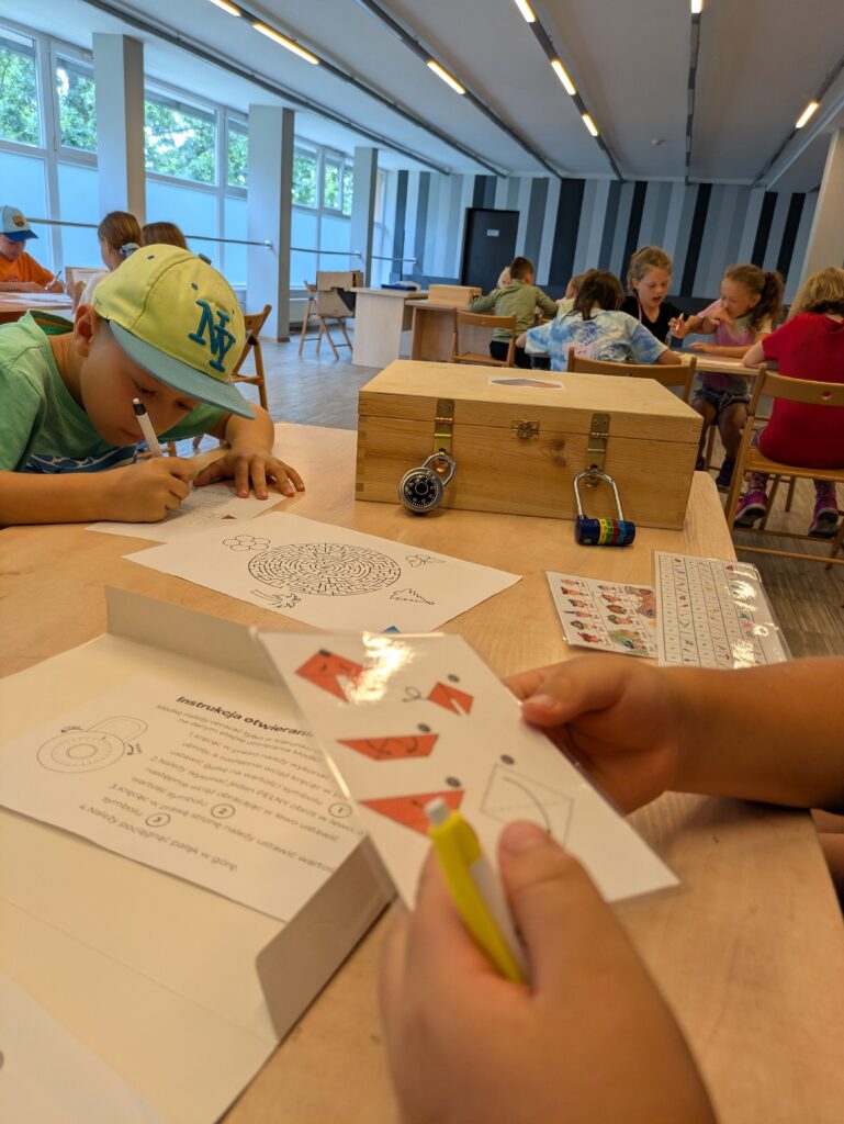Mobilna gra logiczna dla szkół. Uczestnik w czapce z daszkiem rozwiązuje zadanie na kartce obok zamkniętego escape boxa z kłódkami.