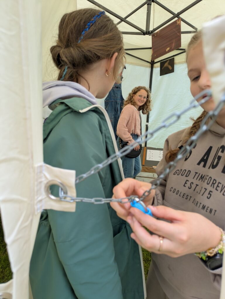 Nastolatki rozwiązujące zagadki w plenerowym namiocie escape room. Dziewczyny w mobilnym escape roomie na evencie. Uczestniczki współpracują, otwierając łańcuch i rozwiązując łamigłówki w namiocie.