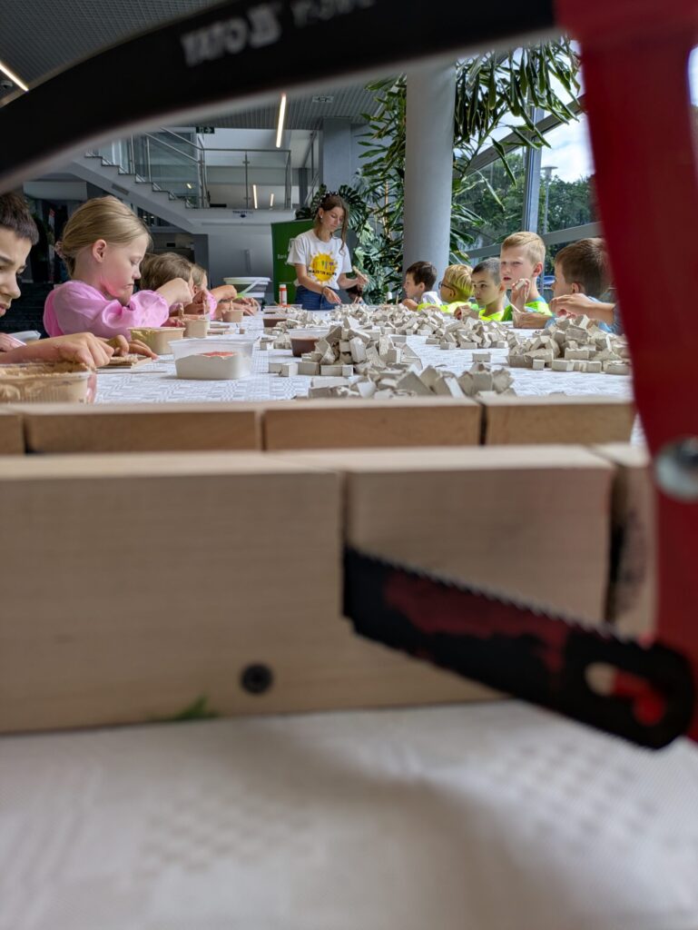 Dzieci siedzące przy długim stole podczas warsztatów budowania miniaturowych domków. Na pierwszym planie widać piłę do drewna.